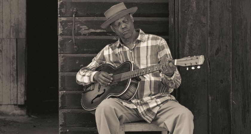 A black-and-white press shot of Eric Bibb playing some acoustic blues on a chair outdoors.