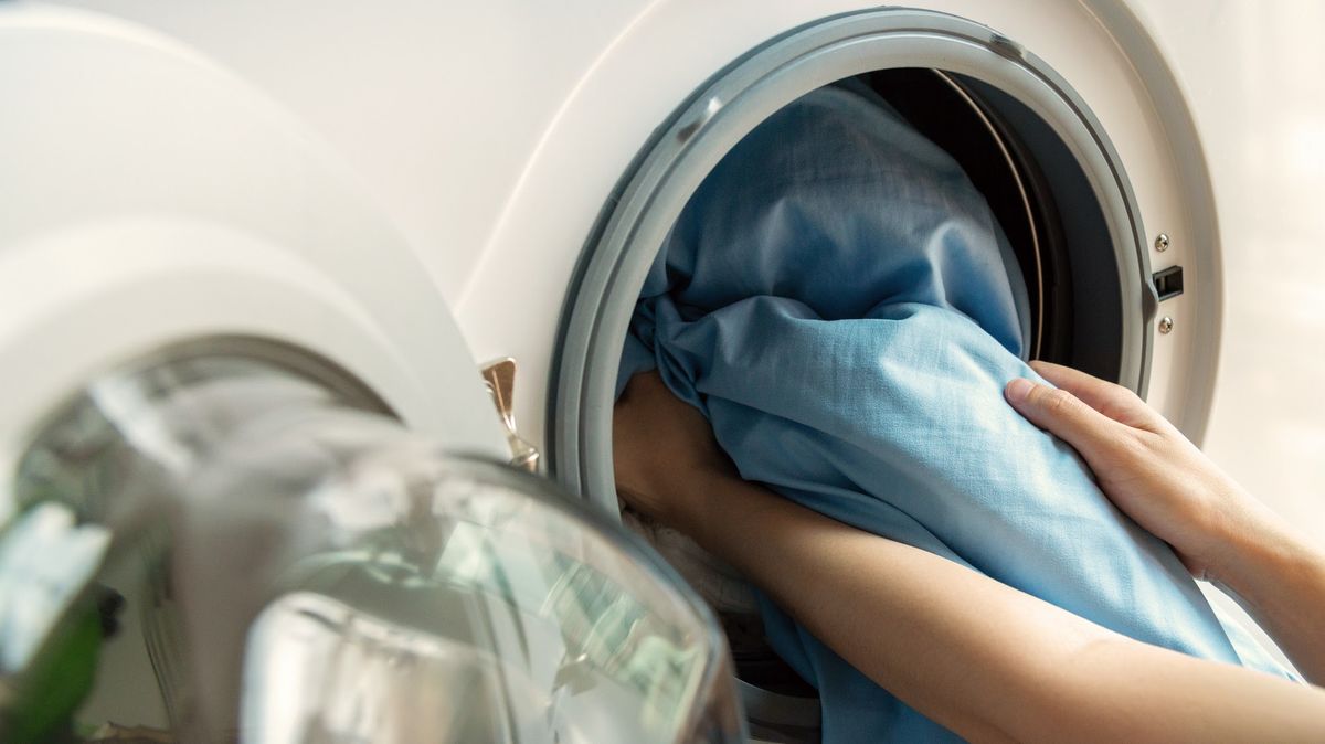 Woman’s hand loading dirty blue bed sheets in a white washing machine.