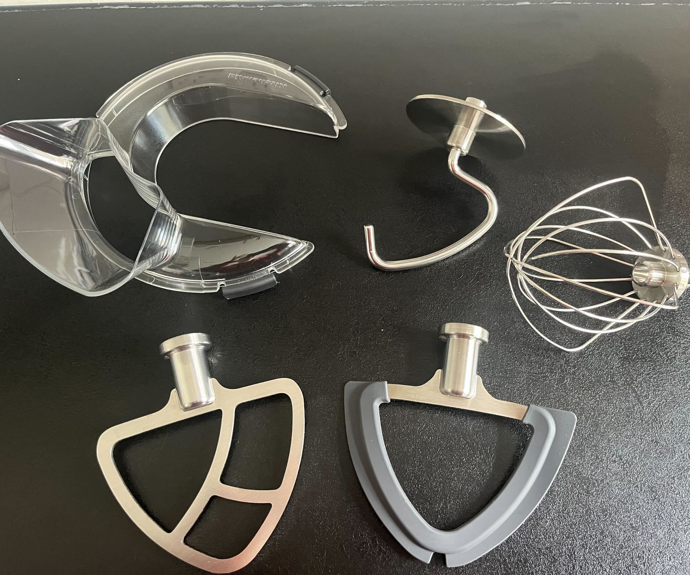 Stainless steel stand mixer attachments laid out on a black counter with a clear plastic spill guard. Two paddles, one whisk, and a dough hook.