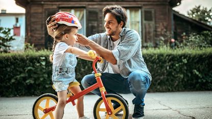Father putting bicycle helmet on toddler daughter on a balance bike