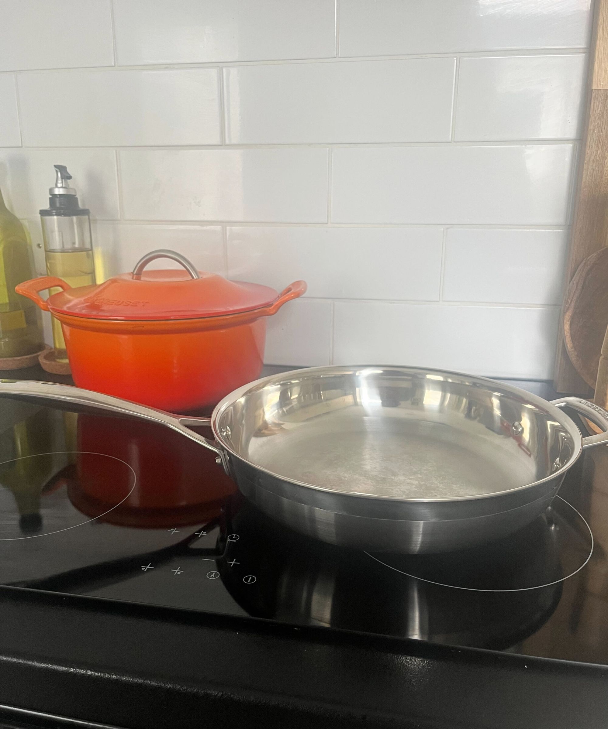 A stainless steel Le Creuset frying pan on a black stovetop, in front of an orange Le Creuset Dutch oven. White tile backsplash behind them.