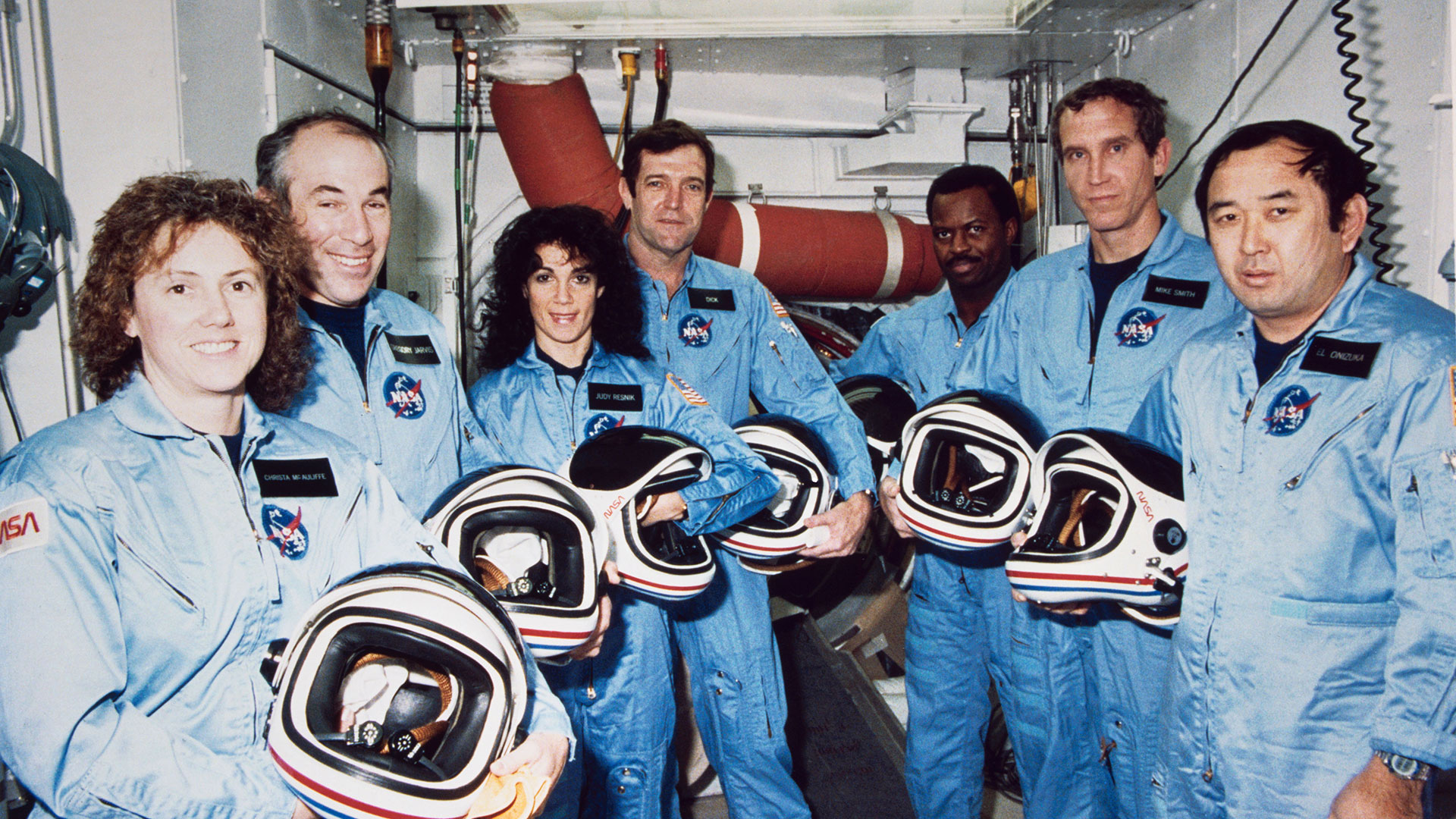 seven people in light blue flight suits and holding white helmets stand in a white-walled room