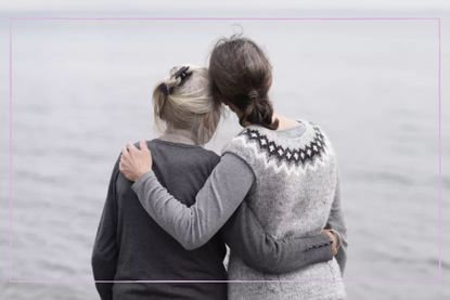 Two women of different ages with grey hair and grey jumpers hugging while looking out over a grey sea.