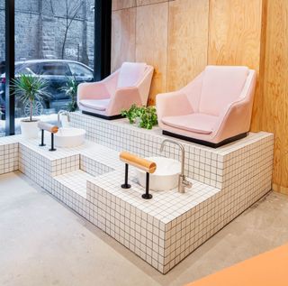 View of the pedicure area at Le Hideout featuring a tiled platform with steps, pink chairs, sinks, taps, wood covered walls, grey floors and a tall window