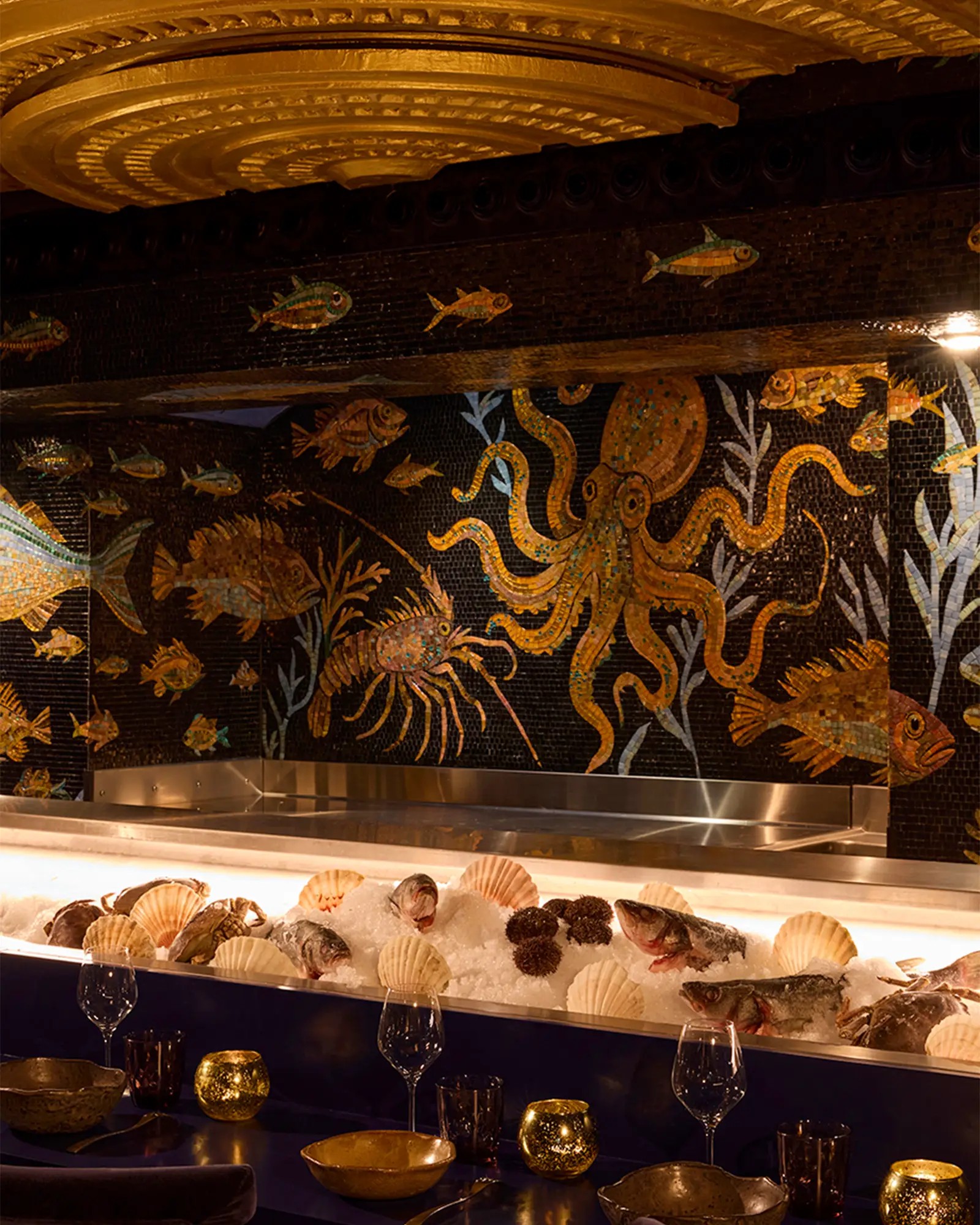 A restaurant mosaiced wall with octopi, fish, and lobsters swimming in a ceramic-clad sea, golden tea light holders, wine glasses, and ceramic bowls for plates.
