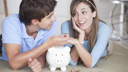 A young couple smile at each other sitting in front of a piggybank while the man holds some pennies.