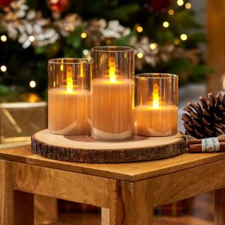 Three flameless candles in clear glass votives on a wooden table with a lit tree in the background.