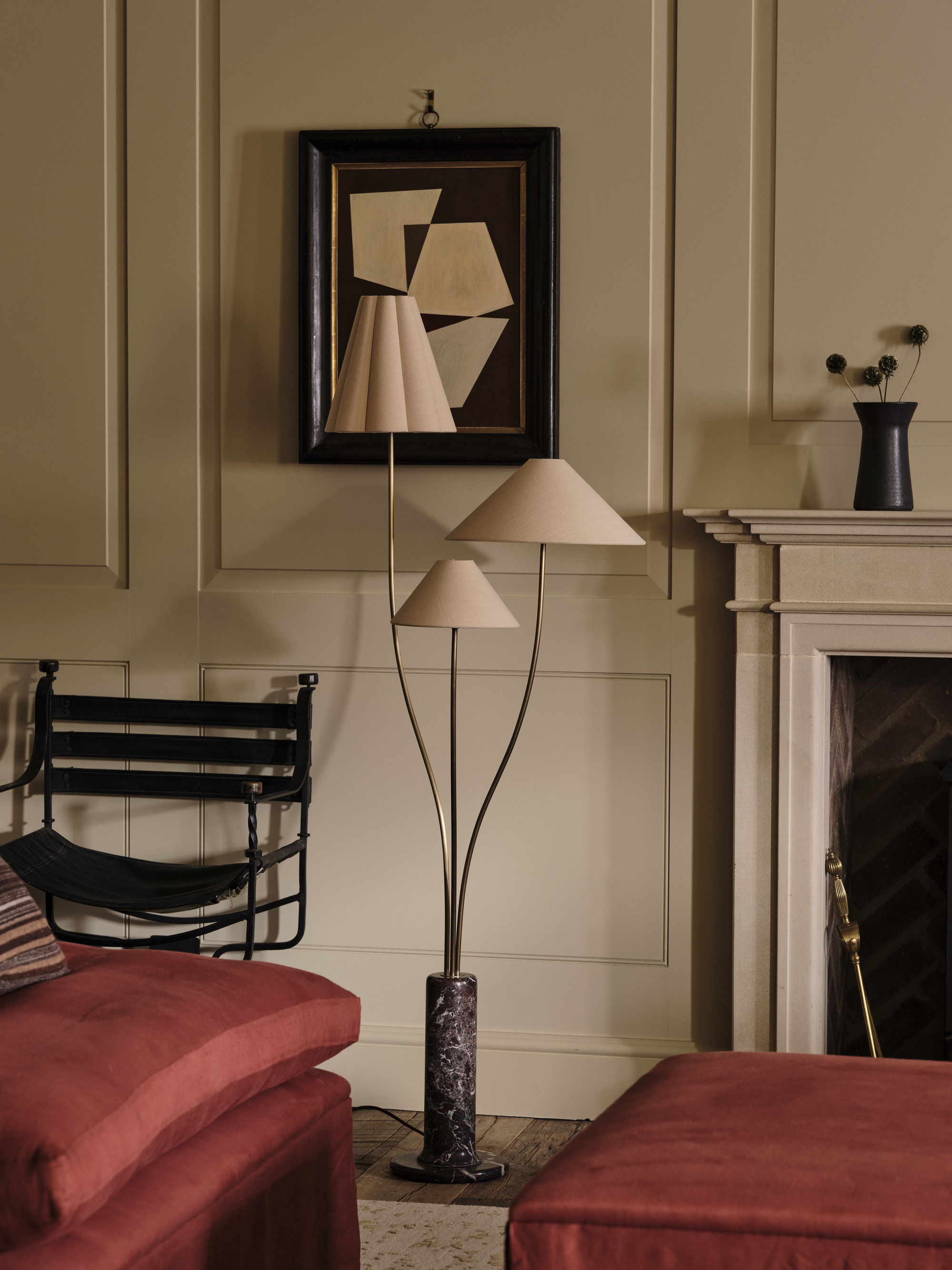 Image of a three-light floor lamp with a marble base in an off-white living room with a photo on the wall, a fireplace, red seating, and a black accent chair.