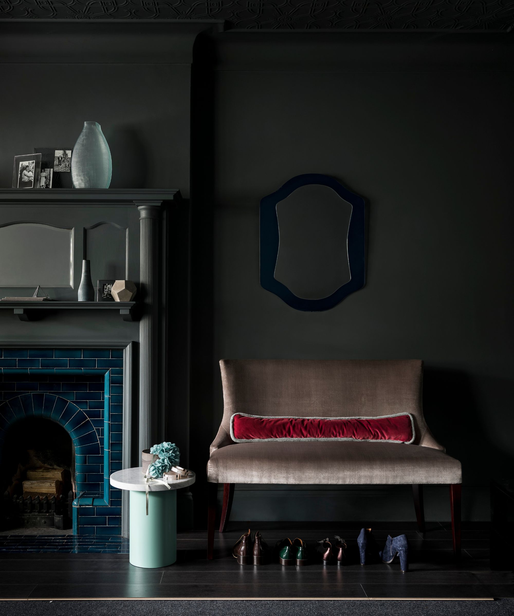 A dark living room with dark greeny grey walls, a blue fireplace with matching dark mantelpiece, a large beige upholstered chair with a row of shoes underneath, and a green circular table with white top to the left of it.
