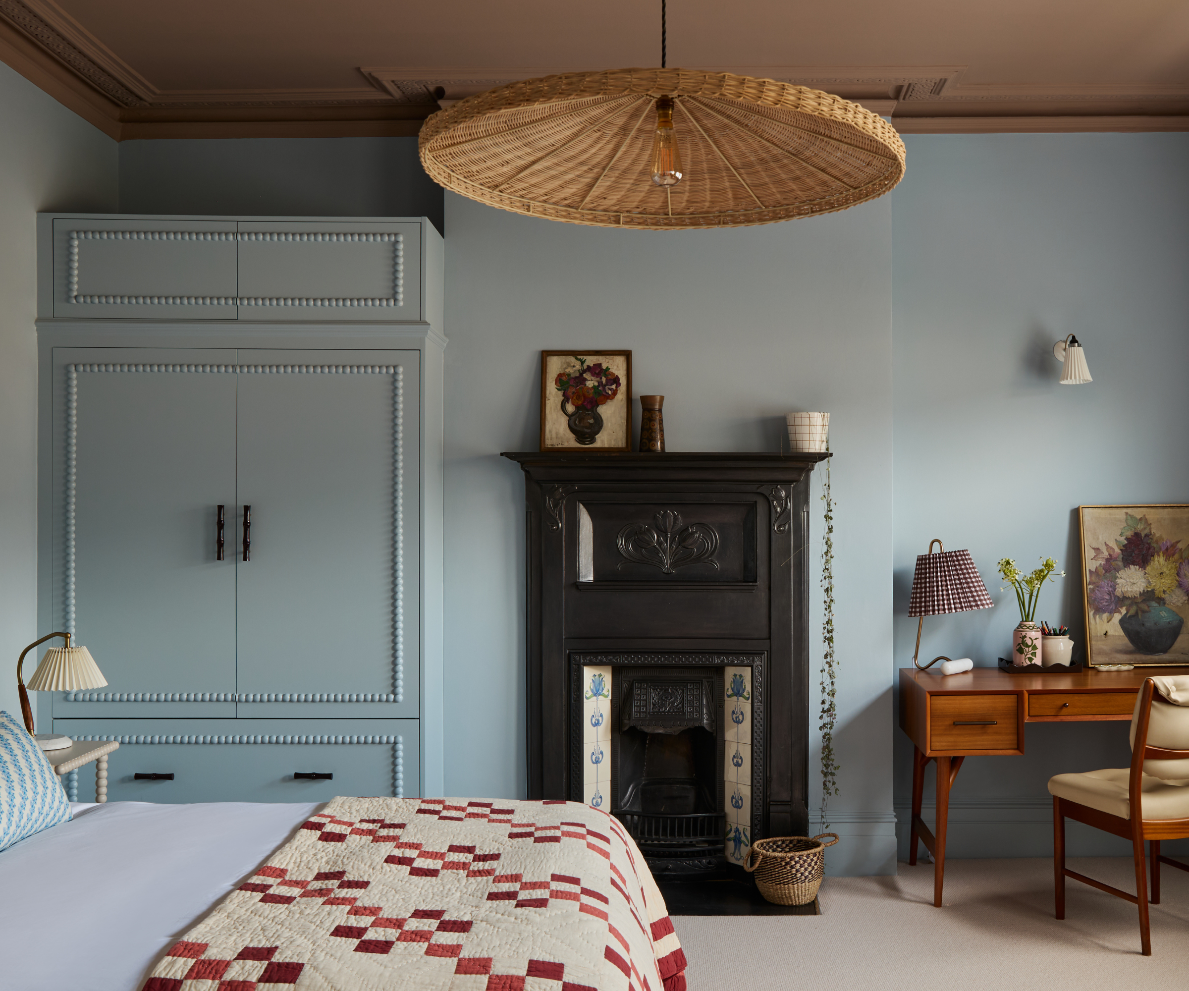 Bedroom with powder blue walls, salmon pink ceiling, rattan pendant light, bed with vintage red and cream quilt, victorian fireplace, wooden desk and a built-in wardrobe painted blue