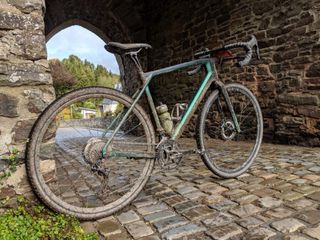 This is a picture of a muddy gravel bike shown side on leaning up against a stone bridge with a cobbled road underneath. The handle bars are pointing away to the right, with the rear wheel left of image slightly closer to the camara.