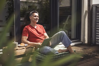 Man relaxing on balcony with digital tablet in hands. He appears to be in his 50s.
