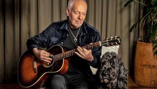 Peter Frampton plays an acoustic guitar in a chair as his service dog Bigsby sits to his right. 