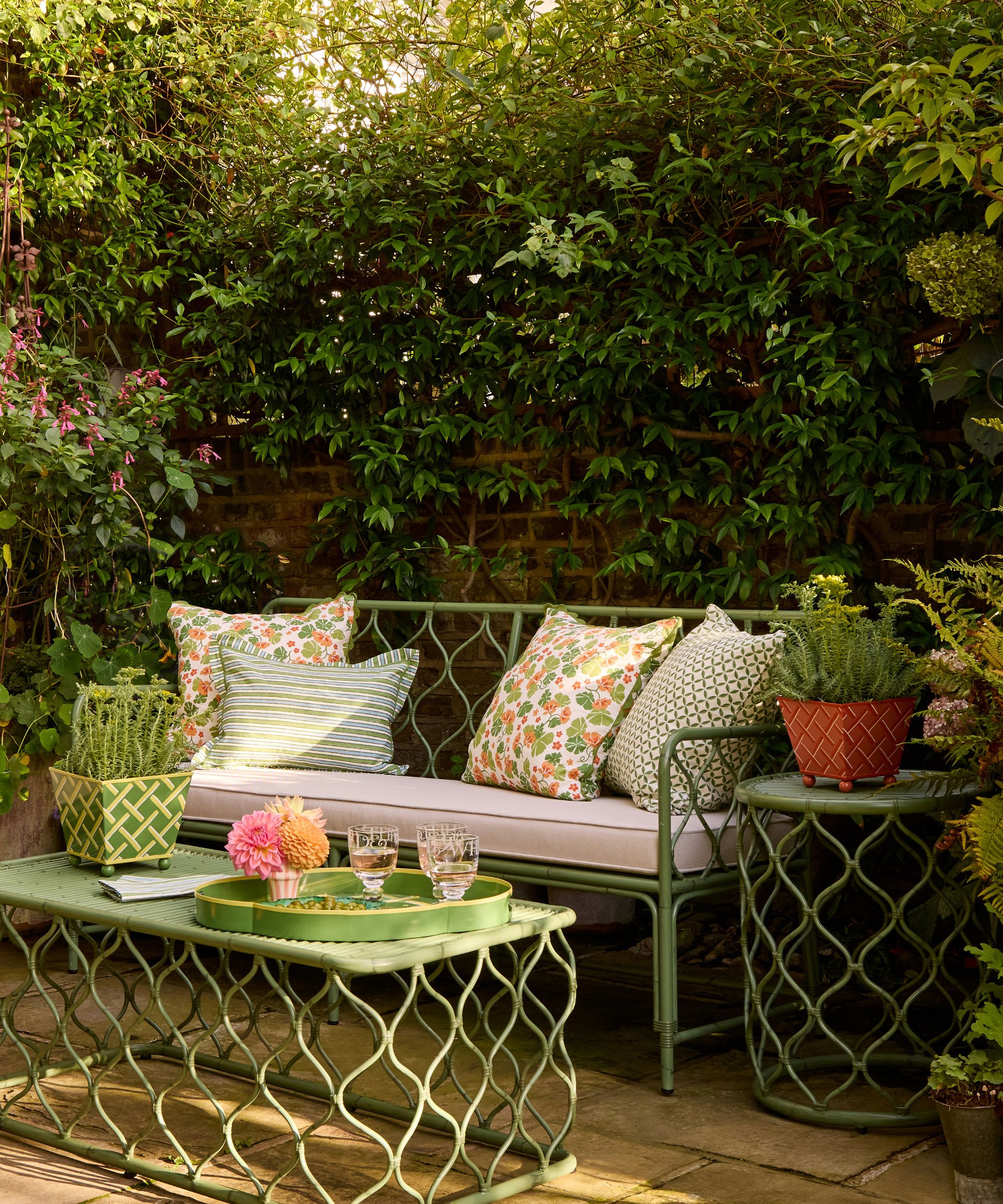 green trellis style rattan outdoor furniture on a small walled garden patio with floral pilows and a coffee table styled with a potted plant and drinks tray