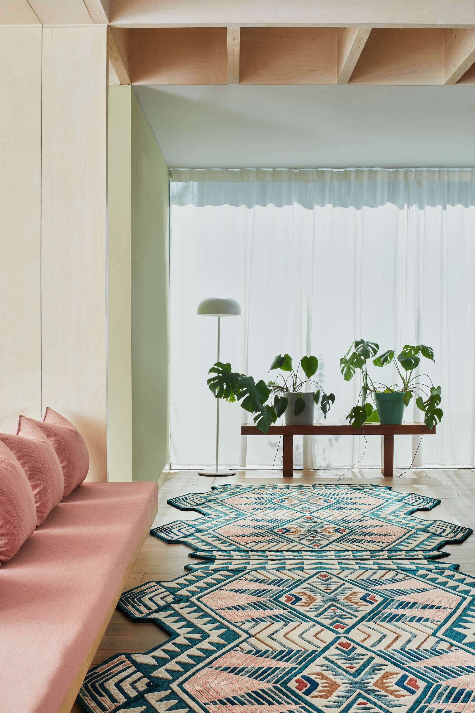 A light living space with a blush pink bench seat to the left and an Aztec style rug in front, and floor to ceiling, curtained windows and the back of the space.