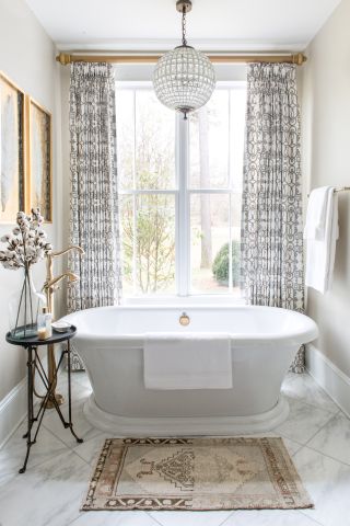 neutral bathroom with white roll top bath, chandelier, marble floor tiles black and white curtains