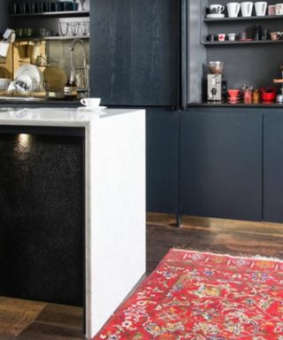 A kitchen island with a white waterfall and black back panel, on a dark wooden floor with a red rug and with dark blue cabinets with colorful decor on it