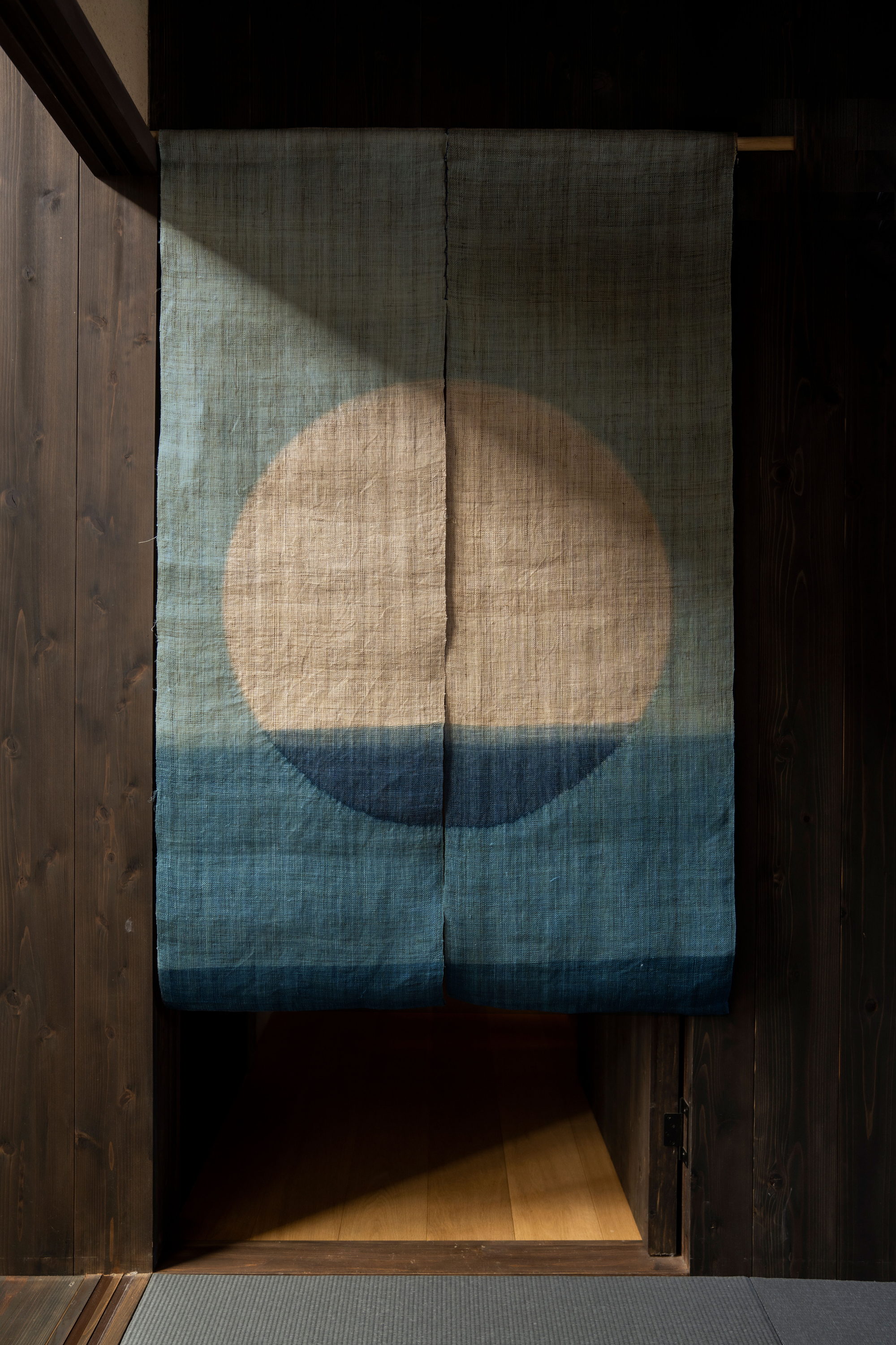Image of blue and teal noren hanging in the doorway of a space with dark wooden walls. There is a shadow across the top of the curtains.