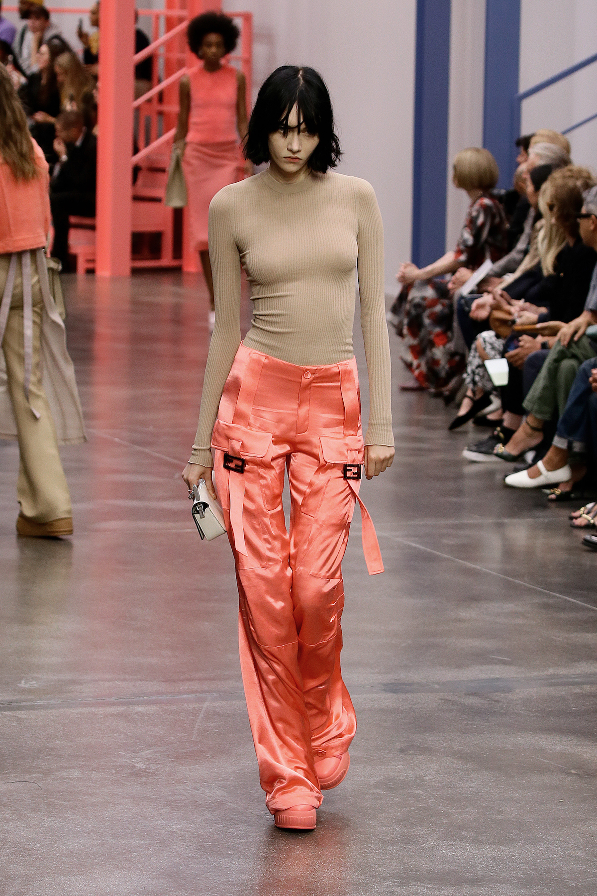 A female model wearing a light brown form fitting long sleeve shirt with shiny pink pants and pink shoes walking down a runway.