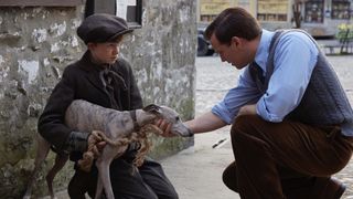 Billy Hickey in a brown cap as Wesley Binks holds onto a grey dog as Nicholas Ralph as James in a blue shirt tends to him in the street in All Creatures Great and Small.