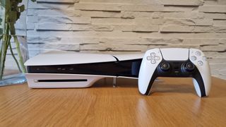 The PS5 Slim and DualSense sitting horizontally sitting on wooden table next to a vase of flowers with a white brick background