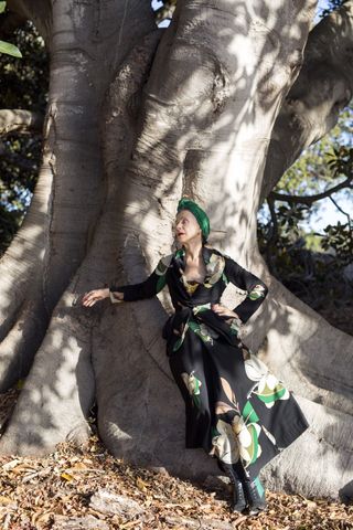 A photo of Beatrix Ost in an over 60 outfit featuring a black and white floral dress with green accents, a green turban, and black lace-up boots leaning against a tree.