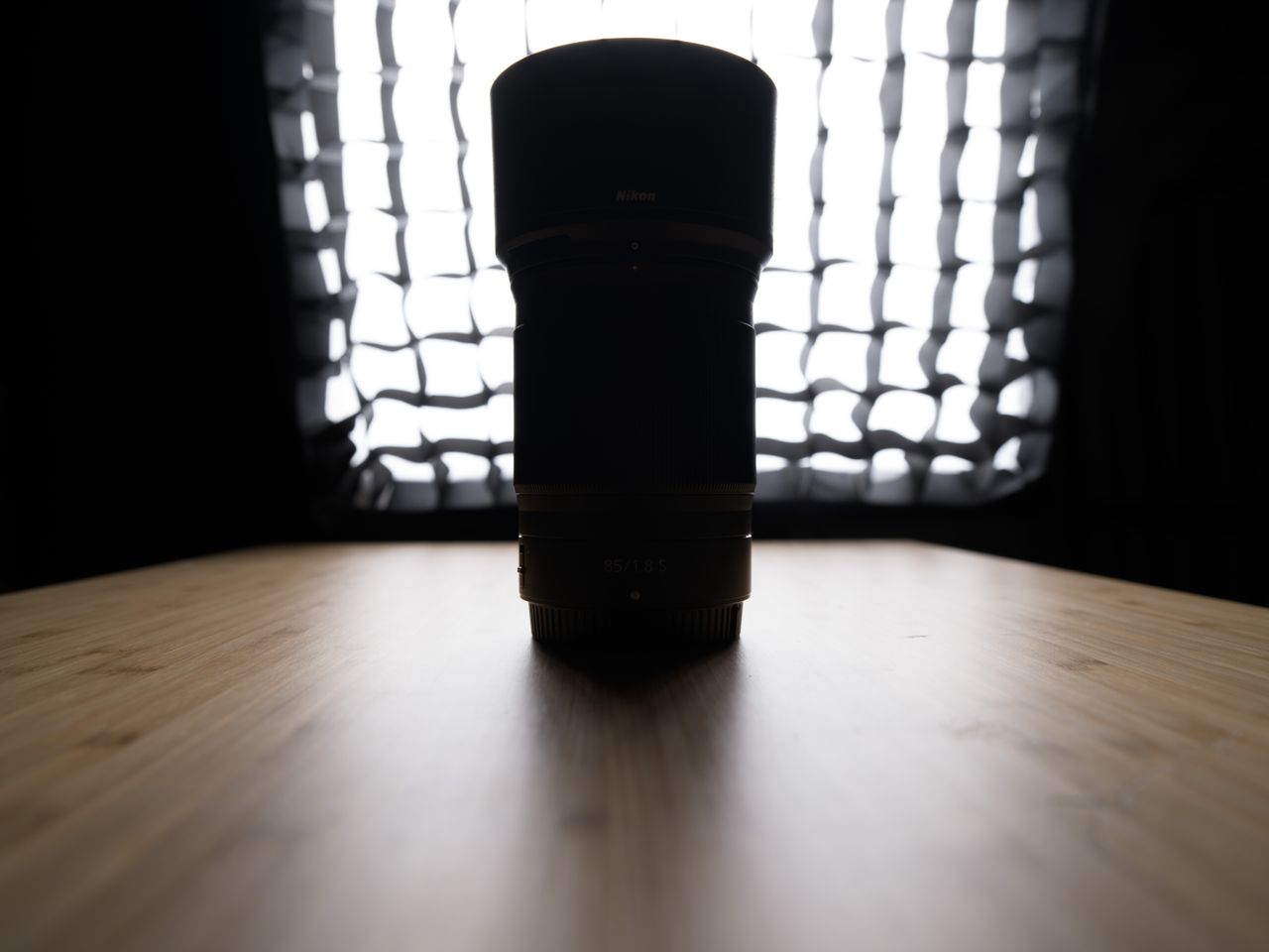 A backlit photo of a Nikon lens on a wooden surface with a studio light in the background. The shadow detail on the lens is not visible, with no post production having been performed.