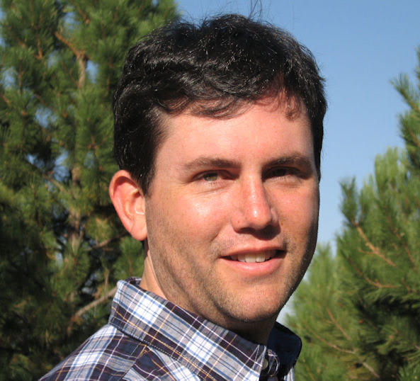 Headshot of man in checked shirt with dark hair in front of pine trees