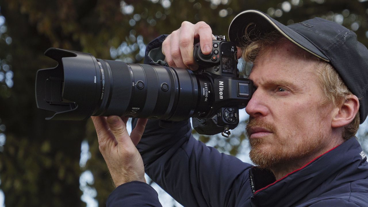 Man holding the Nikon Z8 camera up to his eye and the Nikon Z 70-200mm f/2.8 VR S II lens is attached, there are trees in the background