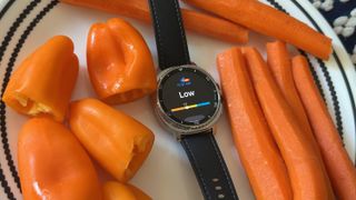 The Samsung Galaxy Watch 8 Classic sitting on a plate surrounded by bell peppers and carrots, the display showing the Antioxidant widget with a "Low" score.