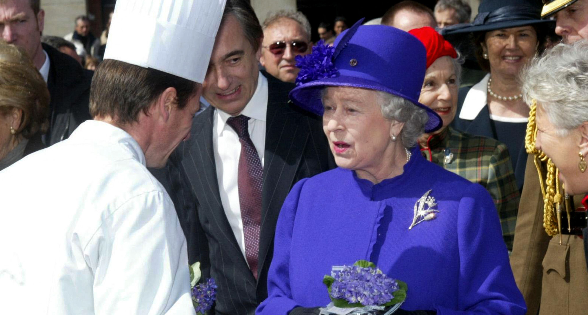 Queen Elizabeth wearing a purple coat and hat speaking to a chef, whose back is to the camera