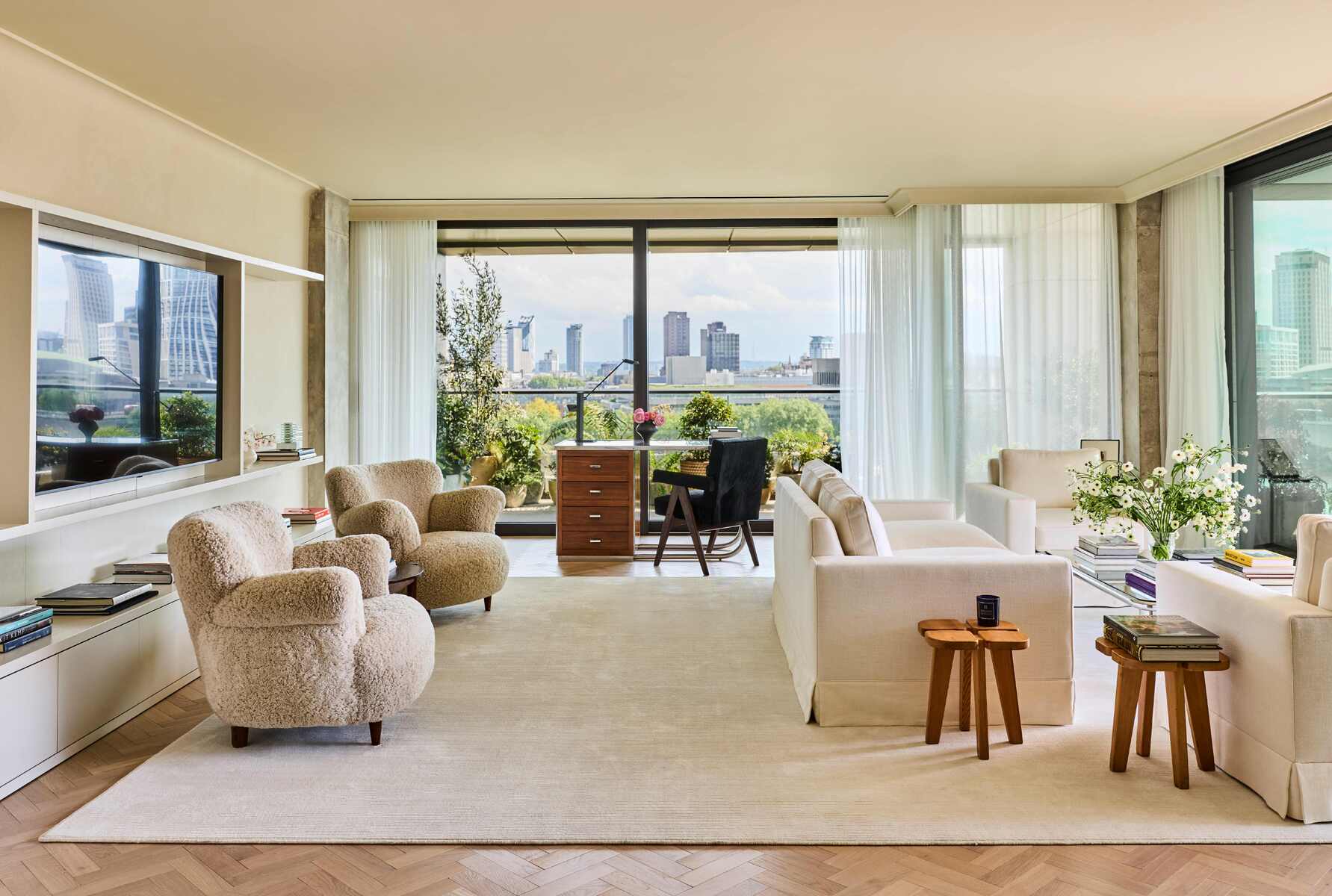 A beige-tinted hotel room with leafy views over a city skyline, shearling armchairs, three sofas, and wooden furniture scattered throughout on wooden flooring and a large carpet.
