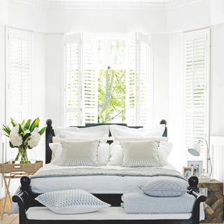 A white bedroom with black double bed and white shutters in a bay window behind