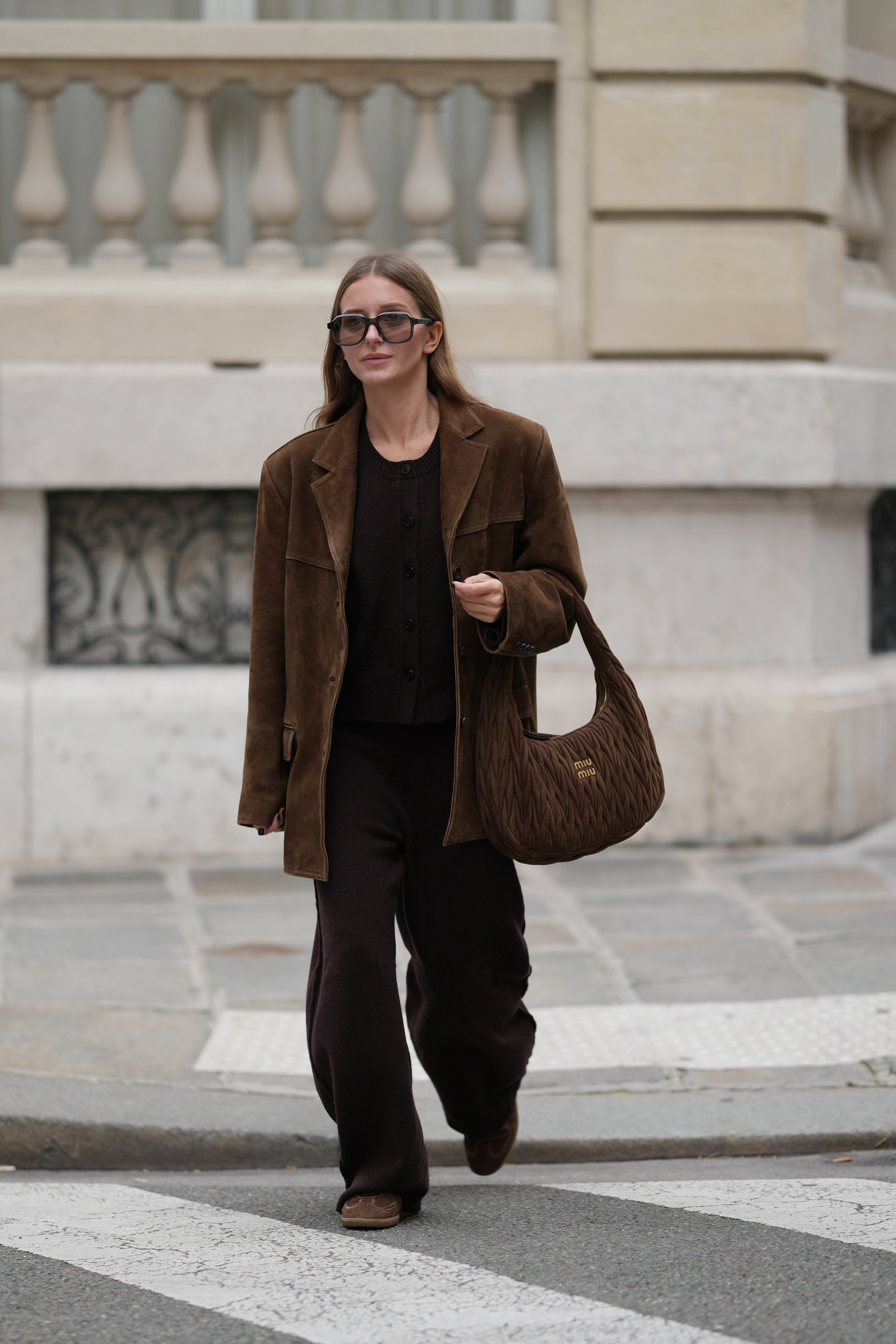 Diane Batoukina wears long straight light brown hair, oversized rectangular sunglasses by Kaleos, a brown suede oversized blazer jacket by Source Unknown, a dark brown knit sweater and matching pants by Dissh, brown suede sneakers by Alohas, and a quilted brown suede Miu Miu bag, during a street style fashion photo session, on October 28, 2025 in Paris, France.