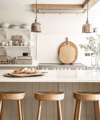 A kitchen island with a marble countertop with wooden chopping boards with two bread loaves on top, three wooden bar stools underneath it, and a kitchen workspace with white decorated shelves, metal lights and an oven