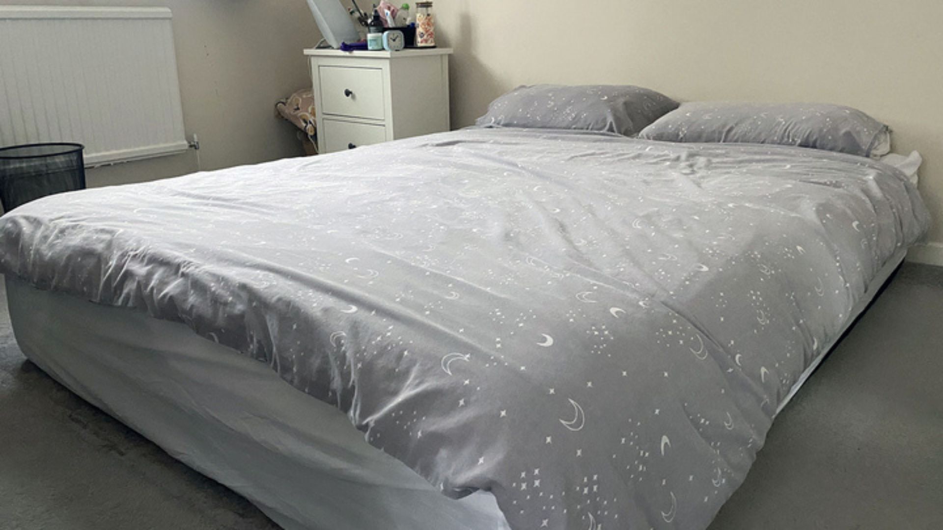 The Nectar Premier Memory Foam Mattress on the floor in out reviewer's bedroom. It's dressed in a gray celestial-print duvet with two pillows resting at the head of the bed. A white bedside cabinet can be seen in the top left-hand corner of the image.