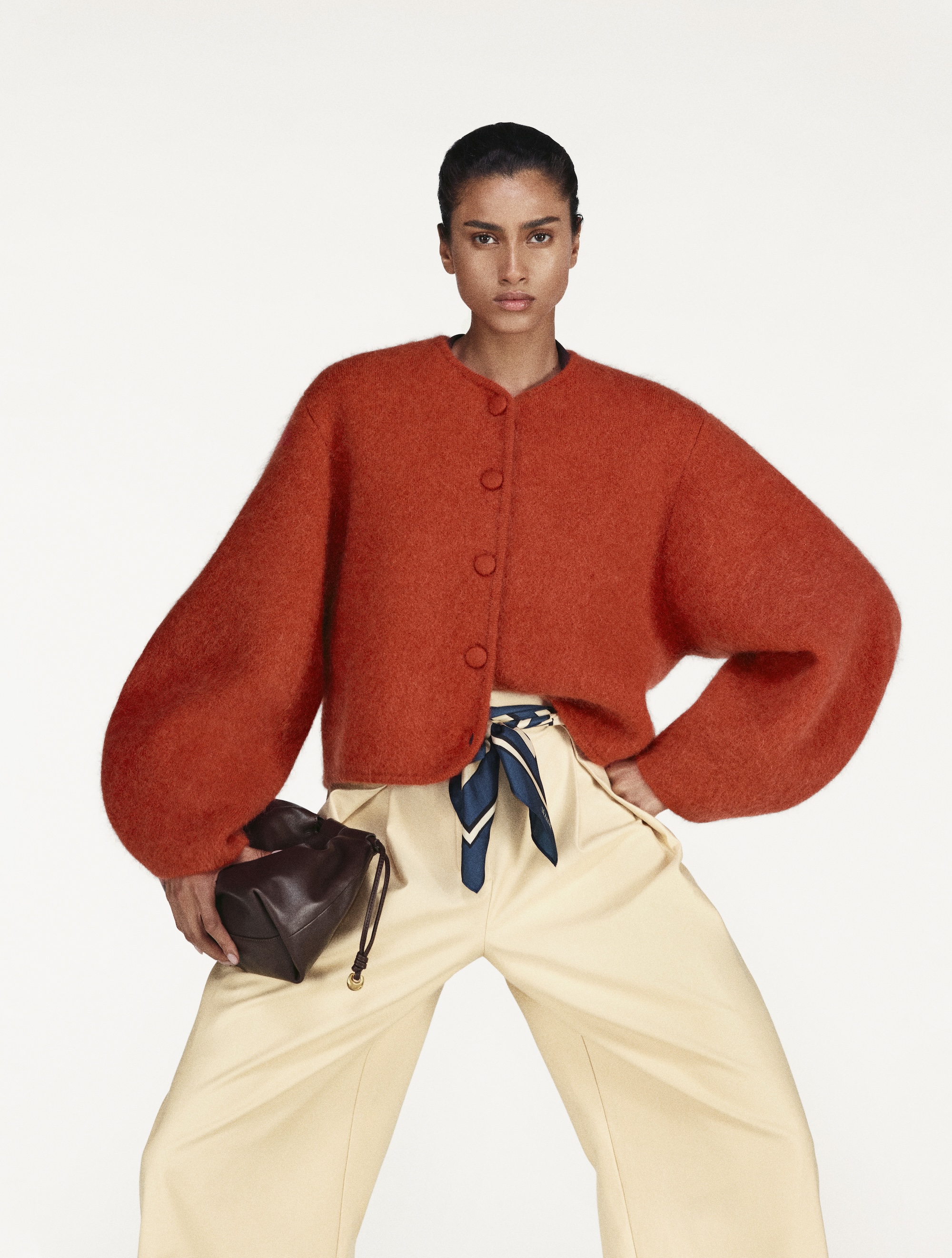 A model wearing an orange-red cardigan with white pants.