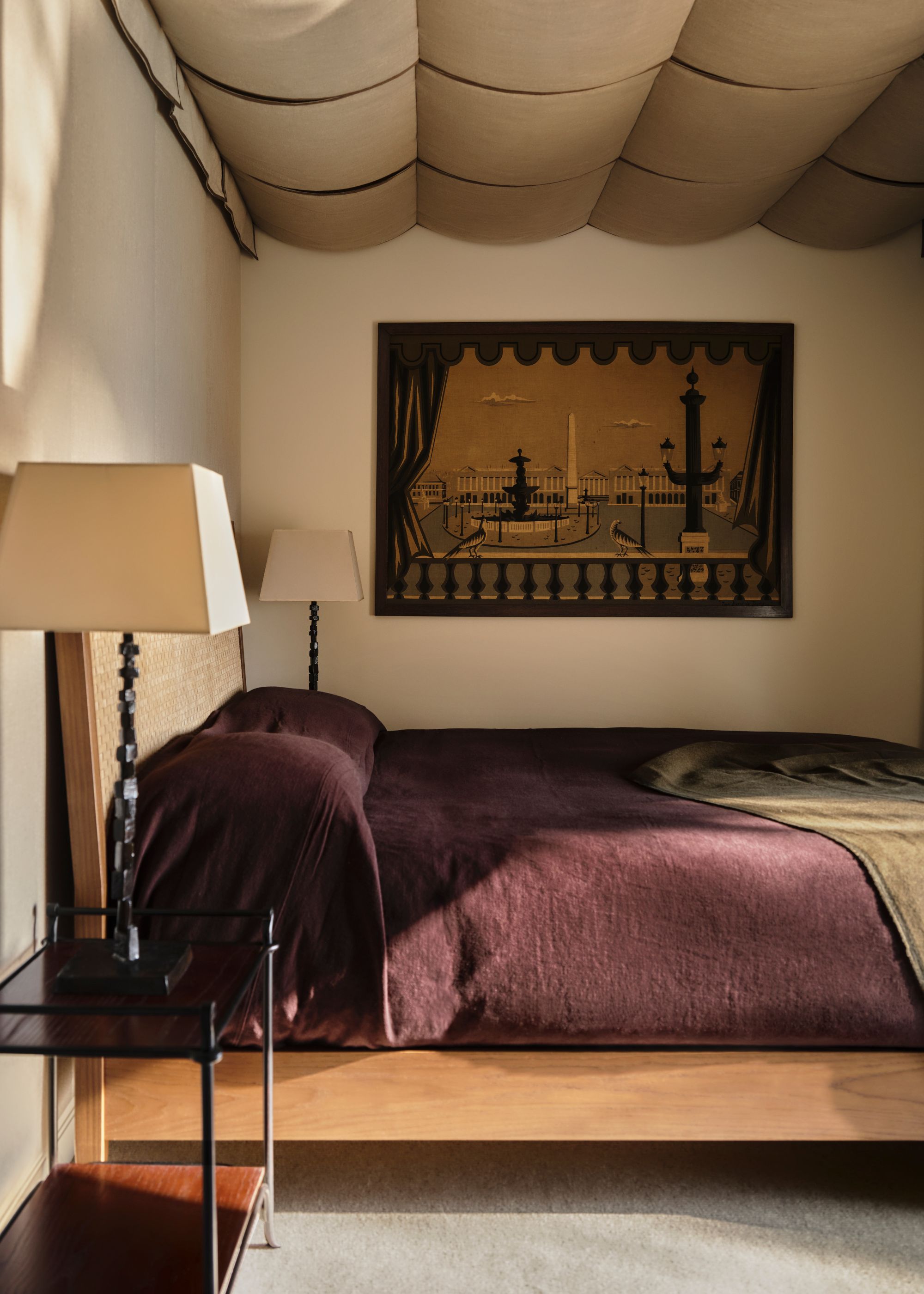 A calming bedroom with beige walls and a draped fabric on the ceiling. A double bed with dark plum linen bedding and neutral, earthy decor.