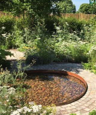 A brown circle water feature with a light brown stone path around it and tall green shrubbery on the borders
