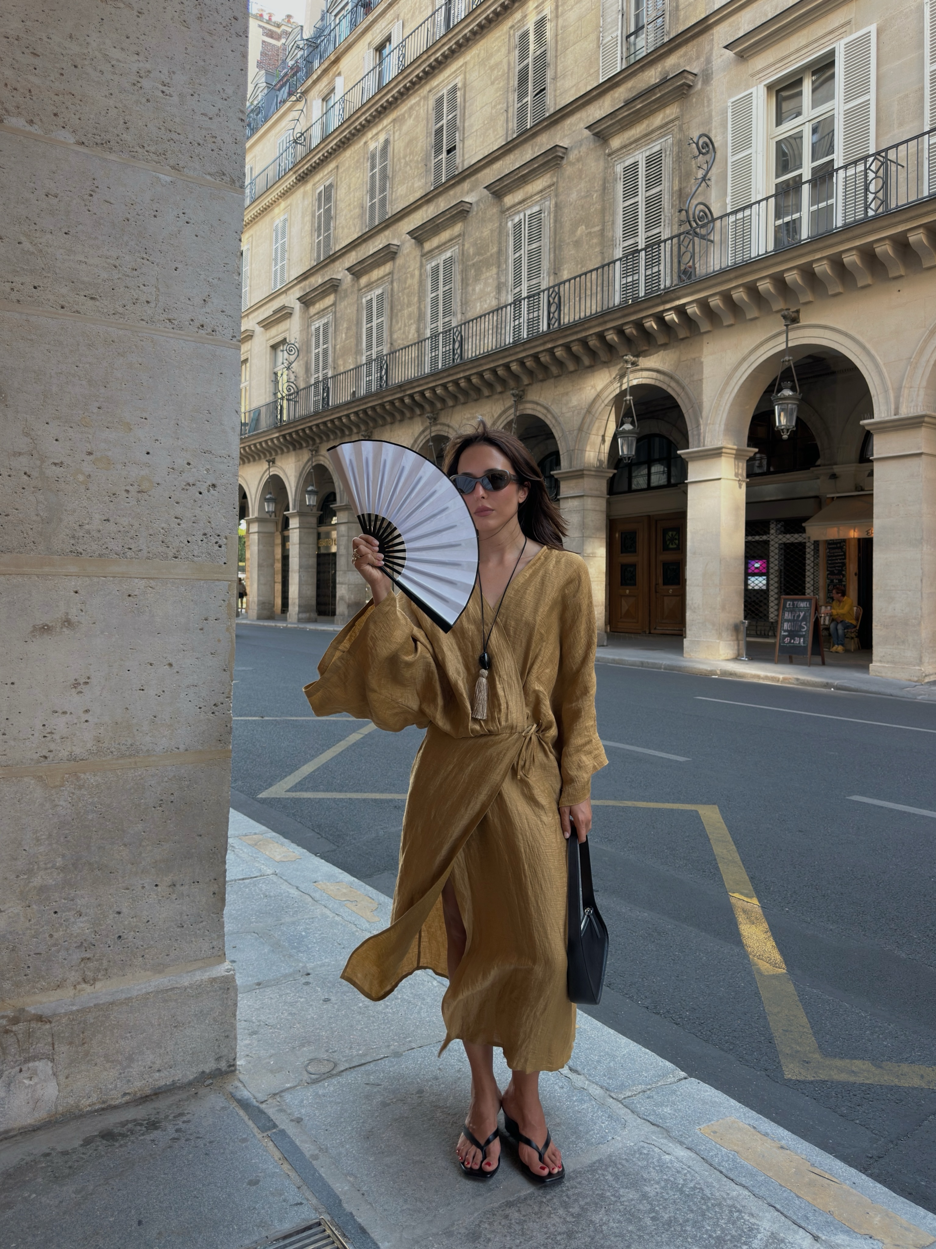An image of an influencer in a yellow wrap dress carrying a white fan and black handbag, wearing black flip-flops and a beige and black tassel necklace in a European city.