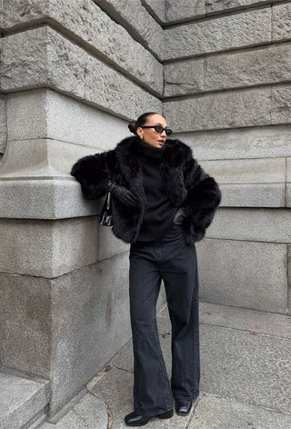 A black turtleneck outfit is shown in a photo of a woman standing on the sidewalk wearing a black fur coat over a black turtleneck with baggy black jeans, black ankle boots, black leather gloves, and a black frame handbag