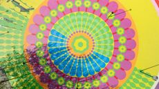 An aerial view of a colorful mural composed of concentric shapes in red, pink, orange, green, and yellow, including flowers, drops, and circles.