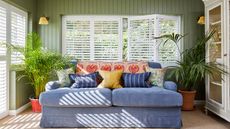 A living room with green shiplap wall panel decor, white vinyl shutter blinds and denim blue upholstered sofa