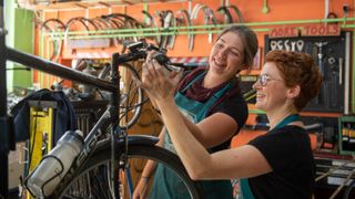 Two women smiling and working together to adjust handlebars in a colourful bike workshop