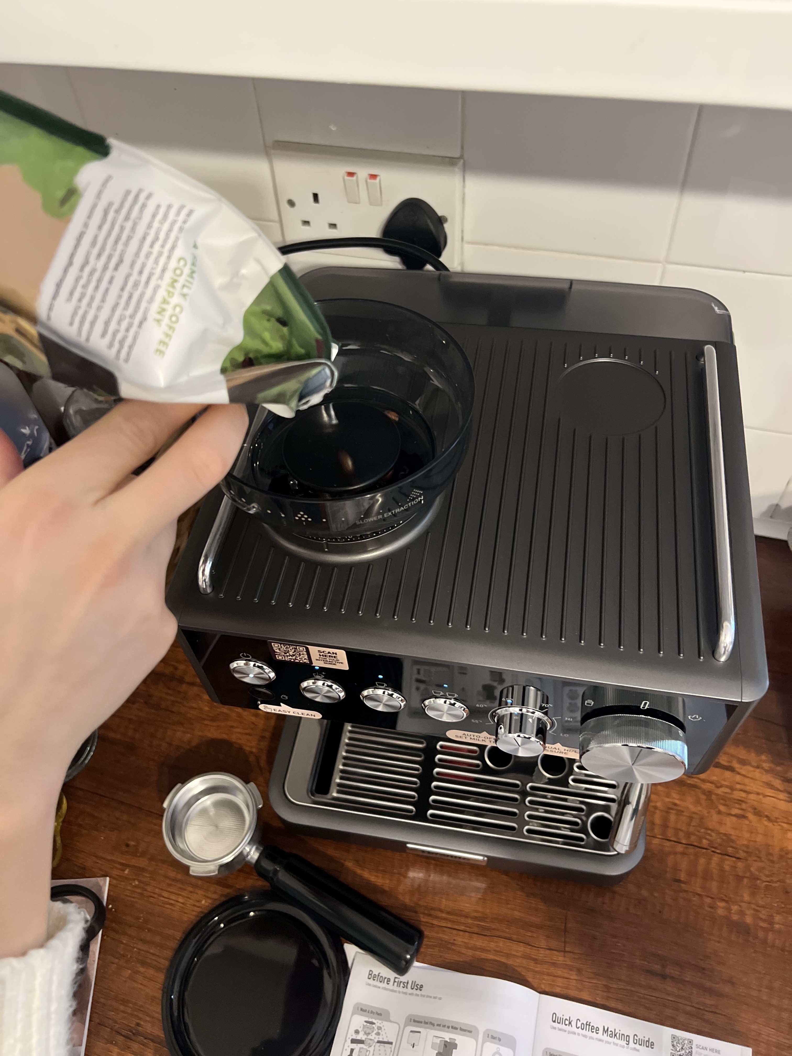 The Breville Barista Sense Espresso Machine in dark gray on a wooden countertop as seen from above. There is a hand pouring coffee beans into the bean hopper.