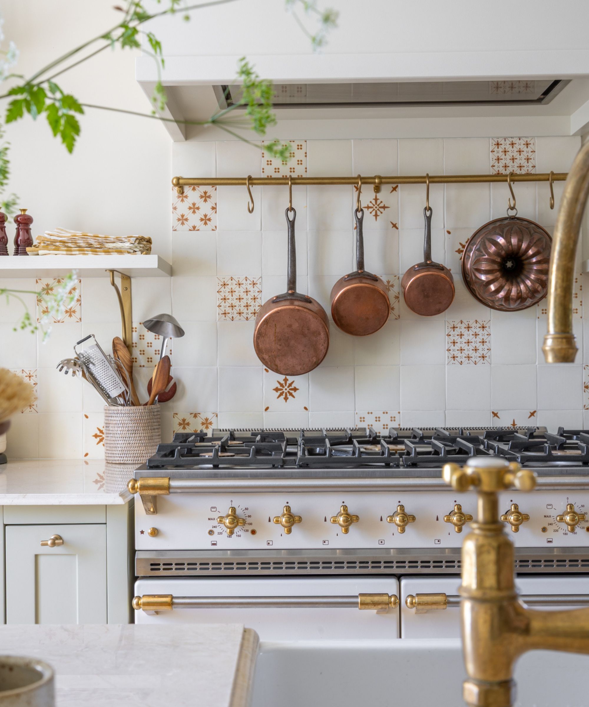 A detailed shot of a white range stove with ornate brass knobs. Above it, three copper saucepans and a copper bundt mold hang from a brass rail against white and terracotta patterned tiles.