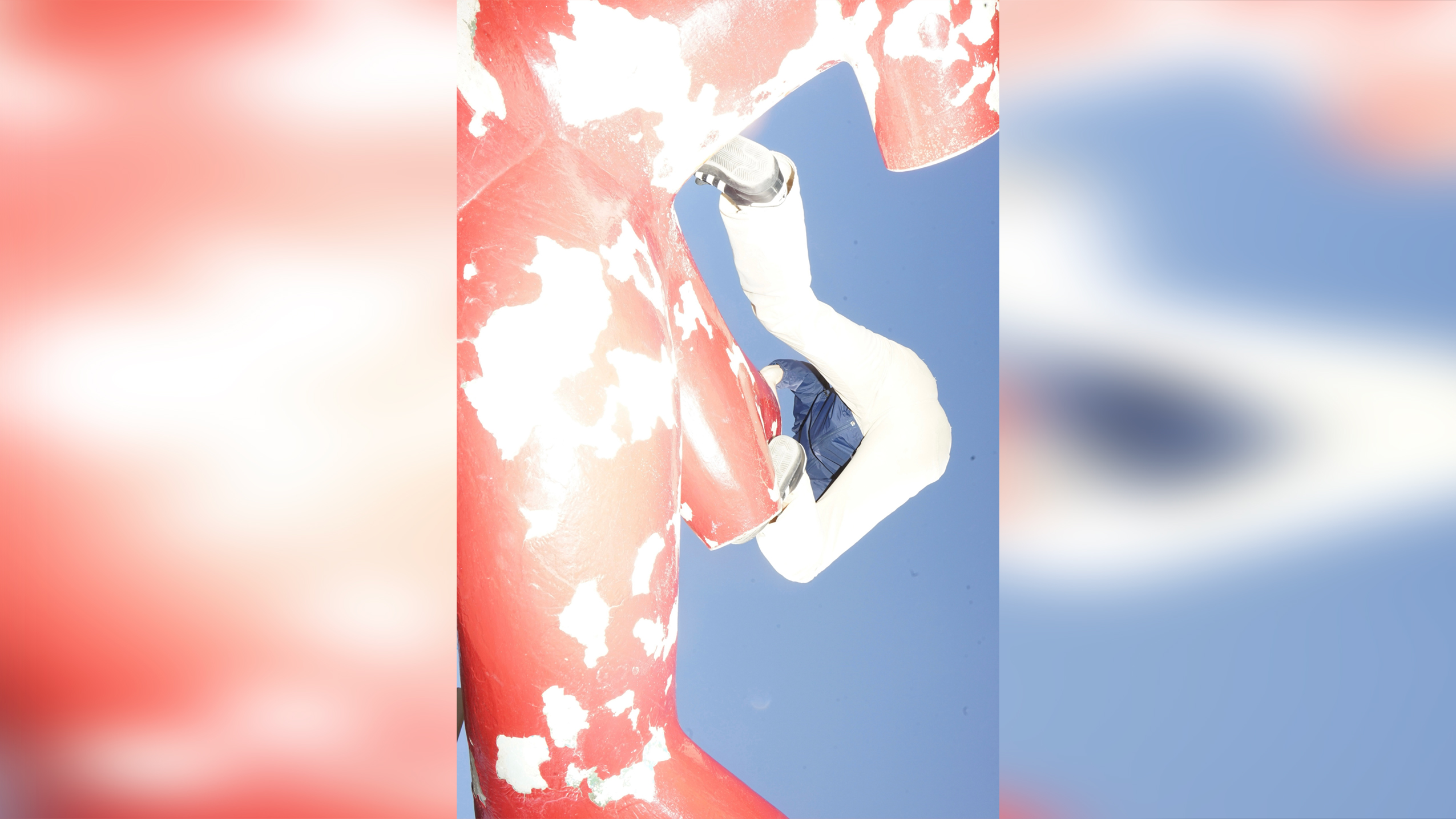 An individual wearing a blue jacket and white pants climbs horizontally across a large, weathered red sculpture with peeling paint against a clear blue sky.