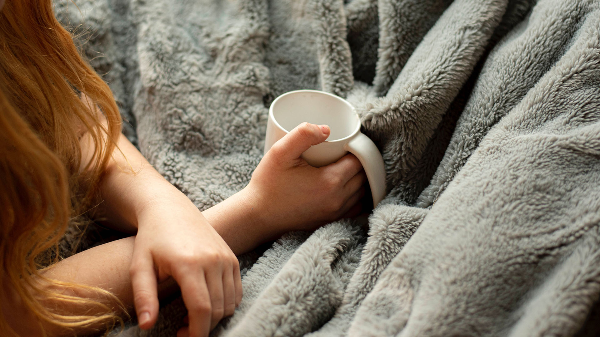 A person holding a mug in bed with a fluffy blanket