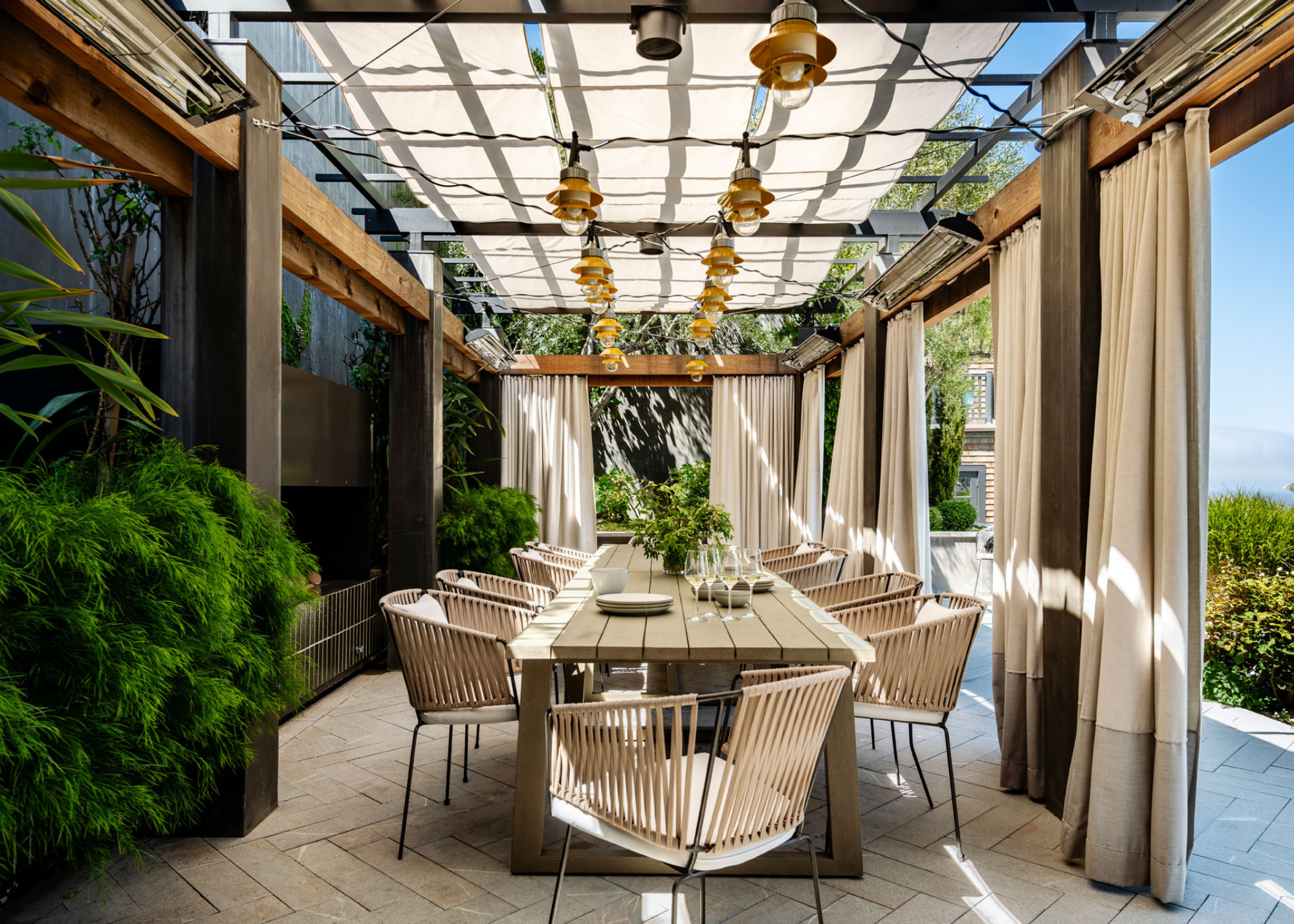 an outdoor dining table underneath a pergola with canopies and lighting overhead and curtains around it