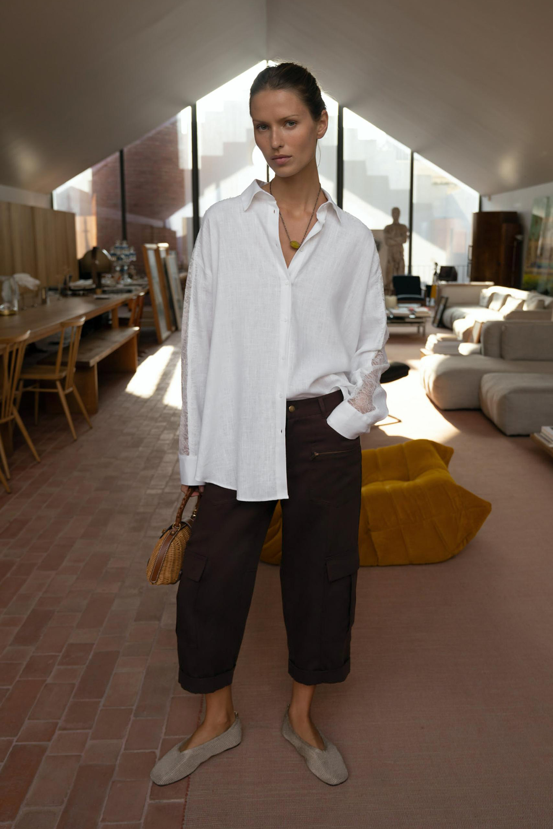 Zara model wearing a pendant necklace, an oversize white button-down shirt, mini wicker basket bag, brown cropped pants, and woven ballet flats.